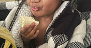 Hailee is registered to the contest to win money with this photo: child, banana, curly_hair, blanket, food, eating, smile, face, hand, indoor, cozy, person, black_and_white, pattern, happy, cute, snack, portrait, expressive, young