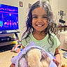 child, girl, smile, curly_hair, toy, container, indoor, living_room, television, tricycle, floor, furniture, happy, person, playful, casual_clothing, home, photography, portrait, cute