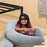 child, smiling, cap, booklet, blue_cushion, carpet, indoor, happy, black_hair, person, blanket, wall, curtain, floor, casual_clothing, young, sitting, cozy, home, relaxed