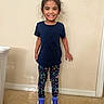 child, girl, smiling, standing, indoor, blue_shirt, patterned_leggings, sneakers, carpet, wall, light_switch, happy, casual_clothing, person, young, cute, portrait, full_body, home, fashion