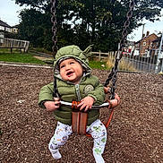 Bowie joined the competition — help win amazing prizes! toddler, child, swing, playground, green_jacket, hood, yoda_ears, smile, happy, outdoor, wood_chips, trees, fence, pants, socks, play, fun, park, daytime, person