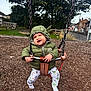 toddler, child, swing, playground, green_jacket, hood, yoda_ears, smile, happy, outdoor, wood_chips, trees, fence, pants, socks, play, fun, park, daytime, person