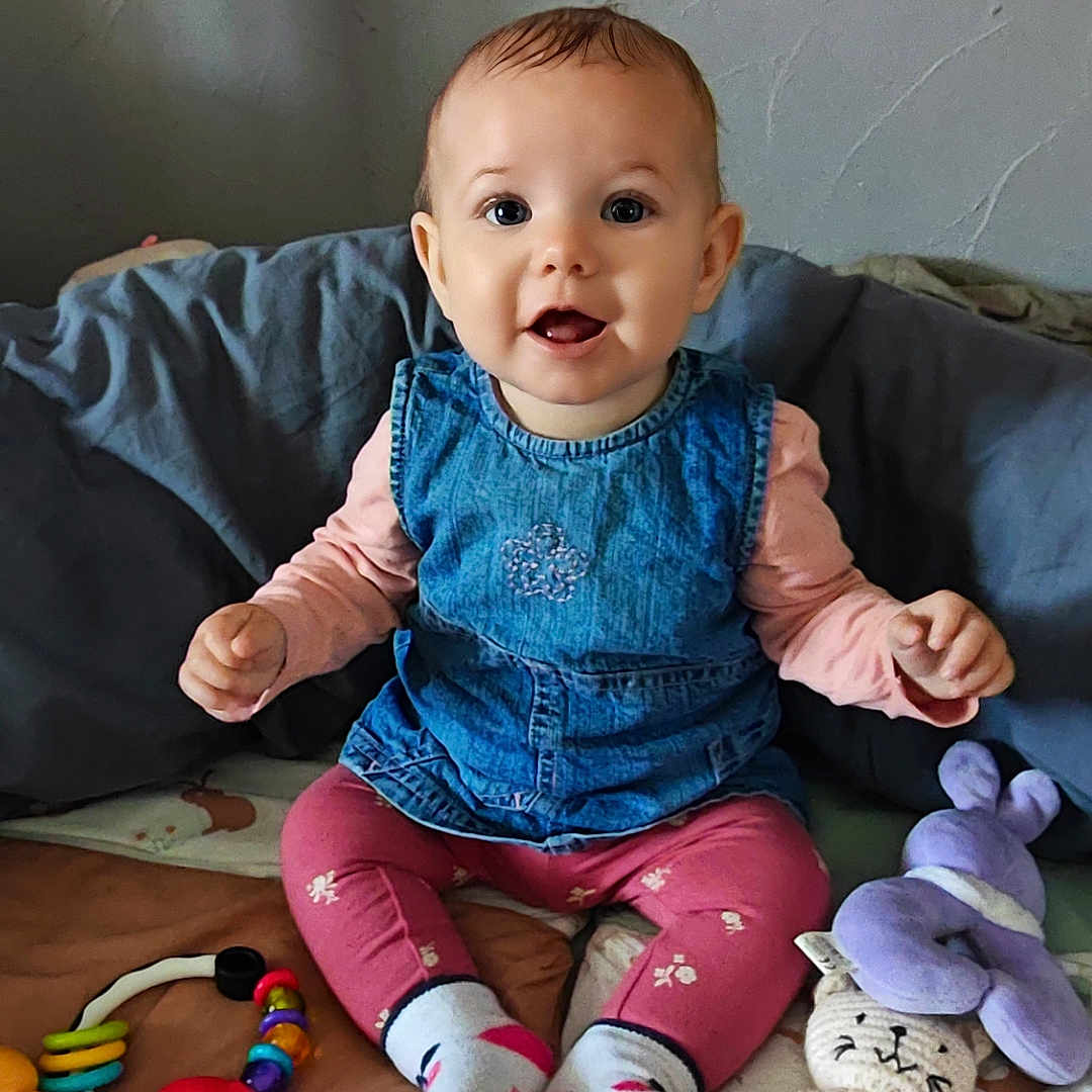 Haïcéa a rejoint le concours — aidez-le/la à gagner de superbes lots ! baby, blanket, child, colorful, cute, denim_dress, face, floor, happy, indoor, person, pink_leggings, playtime, plush_toy, sitting, smiling, socks, teething_ring, toys, wooden_ring