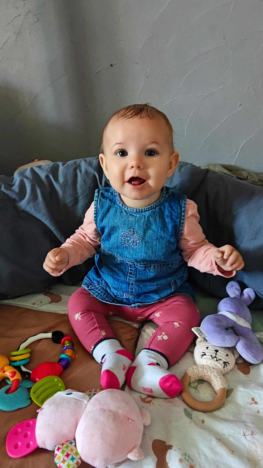 Haïcéa a rejoint le concours — aidez-le/la à gagner de superbes lots ! baby, child, smiling, denim_dress, pink_leggings, socks, toys, plush_toy, teething_ring, colorful, indoor, sitting, happy, person, face, cute, floor, blanket, wooden_ring, playtime