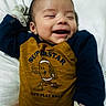 Jonathon is registered to the contest to win money with this photo: baby, infant, child, laughing, smiling, portrait, lying_down, bed, pillow, bedding, indoor, clothing, mustard_outfit, navy_sleeves, striped_socks, cartoon_graphic, text_on_clothing, happy, cozy, close_up