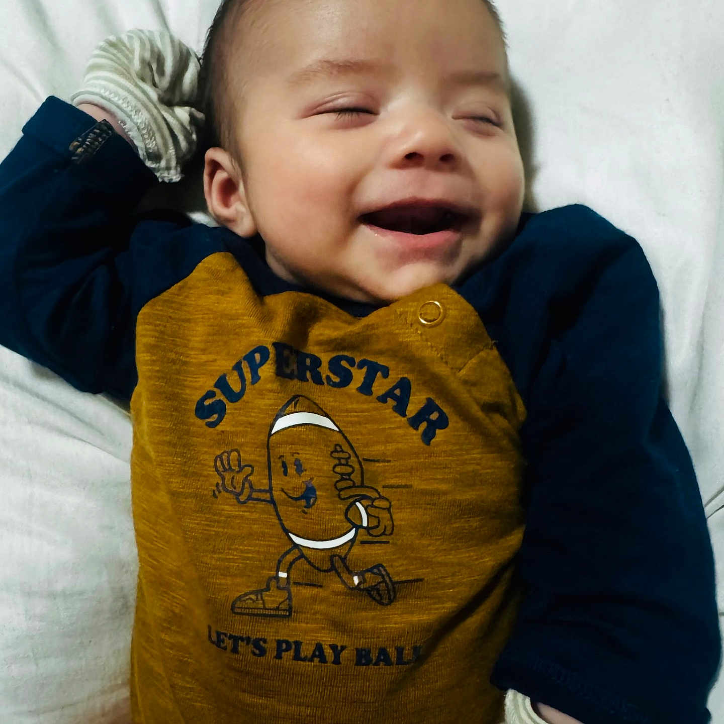 Jonathon is registered to the contest to win money with this photo: baby, infant, child, laughing, smiling, portrait, lying_down, bed, pillow, bedding, indoor, clothing, mustard_outfit, navy_sleeves, striped_socks, cartoon_graphic, text_on_clothing, happy, cozy, close_up