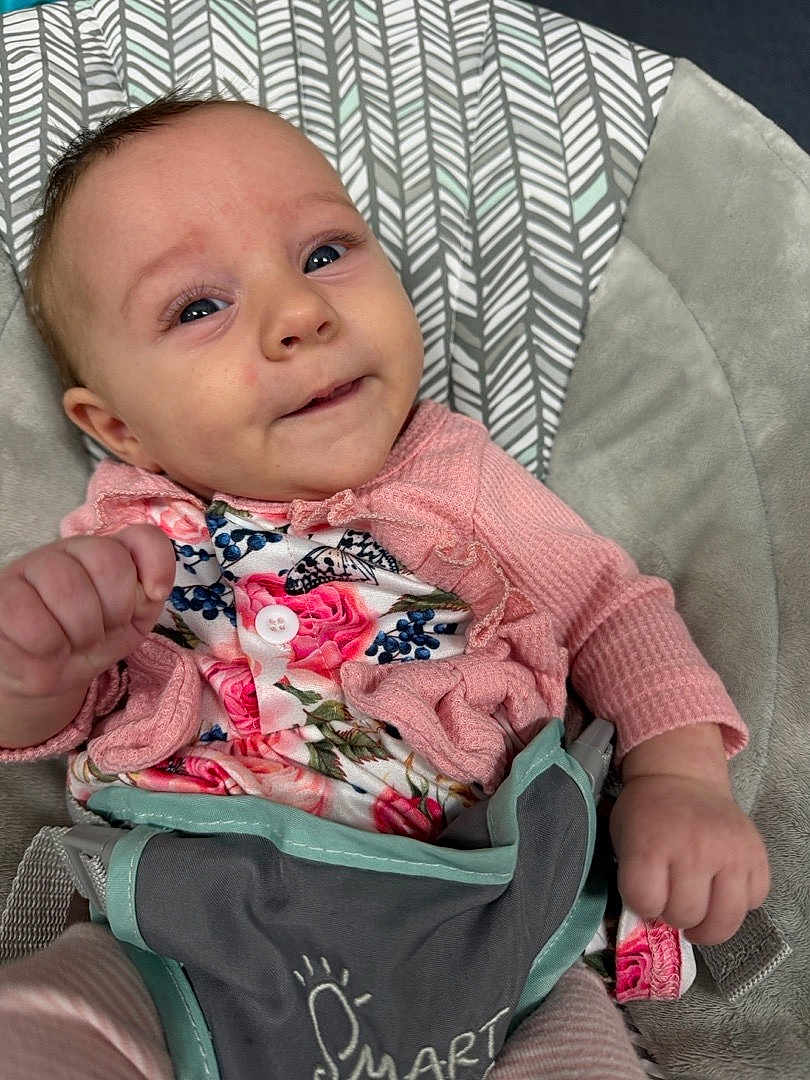 Athena is registered to the contest to win money with this photo: baby, infant, child, pink_clothing, floral_pattern, baby_seat, cushion, face, hand, smile, eyes, cute, portrait, indoors, fabric, pattern, sitting, young_child, soft_texture, comfort