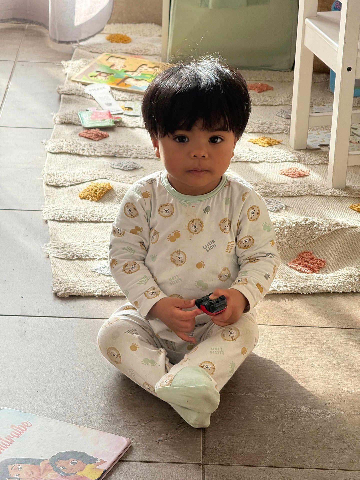 Enaël a rejoint le concours — aidez-le/la à gagner de superbes lots ! toddler, child, toy_car, pajamas, indoor, floor, rug, book, cross_legged, cute, young_child, natural_light, hair, face, expression, play, home, cozy, sitting, person