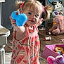 toddler, child, toy, pink_hair_ties, dress, watermelon_pattern, stuffed_animals, blue_toy, floor, wooden_floor, indoor, playtime, curious, pigtails, pink, purple, container, pill_bottle, furniture, candid