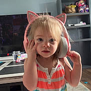 Athena joined the competition — help win amazing prizes! toddler, headphones, cat_ears, child, indoor, striped_dress, blonde_hair, living_room, toy, shelf, television, carpet, furniture, person, cute, curious, colorful_clothing, playful, home, portrait