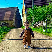 Jules a rejoint le concours — aidez-le/la à gagner de superbes lots ! toddler, child, overalls, plaid_shirt, walking, country_road, stone_buildings, greenery, sunlight, blue_sky, outdoor, daylight, smiling, person, nature, rural, footpath, young_child, casual_clothing, happy