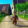 toddler, child, overalls, plaid_shirt, walking, country_road, stone_buildings, greenery, sunlight, blue_sky, outdoor, daylight, smiling, person, nature, rural, footpath, young_child, casual_clothing, happy