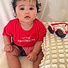Victoria is registered to the contest to win money with this photo: baby, child, red_shirt, little_firecracker, sitting, curly_hair, patriotic_sunglasses, white_pillow, soft_texture, indoor, portrait, cute, infant, expression, face, legs, feet, hand, clothing, background