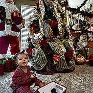 Angel is registered to the contest to win money with this photo: toddler, child, christmas_tree, holiday_decorations, santa_figure, reindeer_decoration, carpet, plaid_pajamas, remote_control, gift_box, indoors, window_blinds, garland, stairs, festive, smiling, holiday_lights, red_clothing, decorative_ribbons, christmas_ornaments