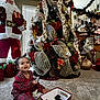 toddler, child, christmas_tree, holiday_decorations, santa_figure, reindeer_decoration, carpet, plaid_pajamas, remote_control, gift_box, indoors, window_blinds, garland, stairs, festive, smiling, holiday_lights, red_clothing, decorative_ribbons, christmas_ornaments