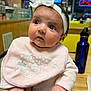 baby, infant, bib, headband, bow, big_eyes, curious_expression, person, portrait, close_up, hand, wooden_table, water_bottle, bottle, restaurant, indoor, clothing, skin, table, cute