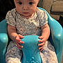 baby, child, infant, blue_chair, floral_outfit, socks, hands, face, eyes, living_room, couch, cushion, carpet, indoor, seated, curious_expression, molded_chair, person, cute, young_child