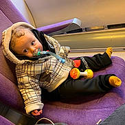 Lino participe au concours pour gagner de l'argent avec cette photo : baby, child, pacifier, jacket, toy, train_seat, purple_seat, window, curly_hair, clothing, socks, indoor, person, seated, infant, cute, relaxed, travel, transport, casual