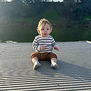 Thiago is registered to the contest to win money with this photo: child, toddler, baby, sitting, lake, dock, outdoors, nature, water, trees, sunlight, backlighting, portrait, face, blond_hair, striped_shirt, shorts, socks, lakeside, daylight