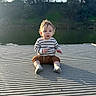 child, toddler, baby, sitting, lake, dock, outdoors, nature, water, trees, sunlight, backlighting, portrait, face, blond_hair, striped_shirt, shorts, socks, lakeside, daylight