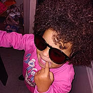 Cordelya a rejoint le concours — aidez-le/la à gagner de superbes lots ! child, kid, sunglasses, pink_sweatshirt, curly_hair, afro, finger_in_mouth, smile, teeth, indoor, flash_photo, candid, playful, portrait, accessory, cluttered_background, doorway, floor_tiles, packaged_goods, person