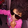 child, kid, sunglasses, pink_sweatshirt, curly_hair, afro, finger_in_mouth, smile, teeth, indoor, flash_photo, candid, playful, portrait, accessory, cluttered_background, doorway, floor_tiles, packaged_goods, person