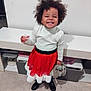 child, smiling, curly_hair, party_hat, red_skirt, white_top, stuffed_animal, toy, boots, indoor, shelf, white_wall, floor, happy, portrait, standing, playful, toddler, clothing, young_child