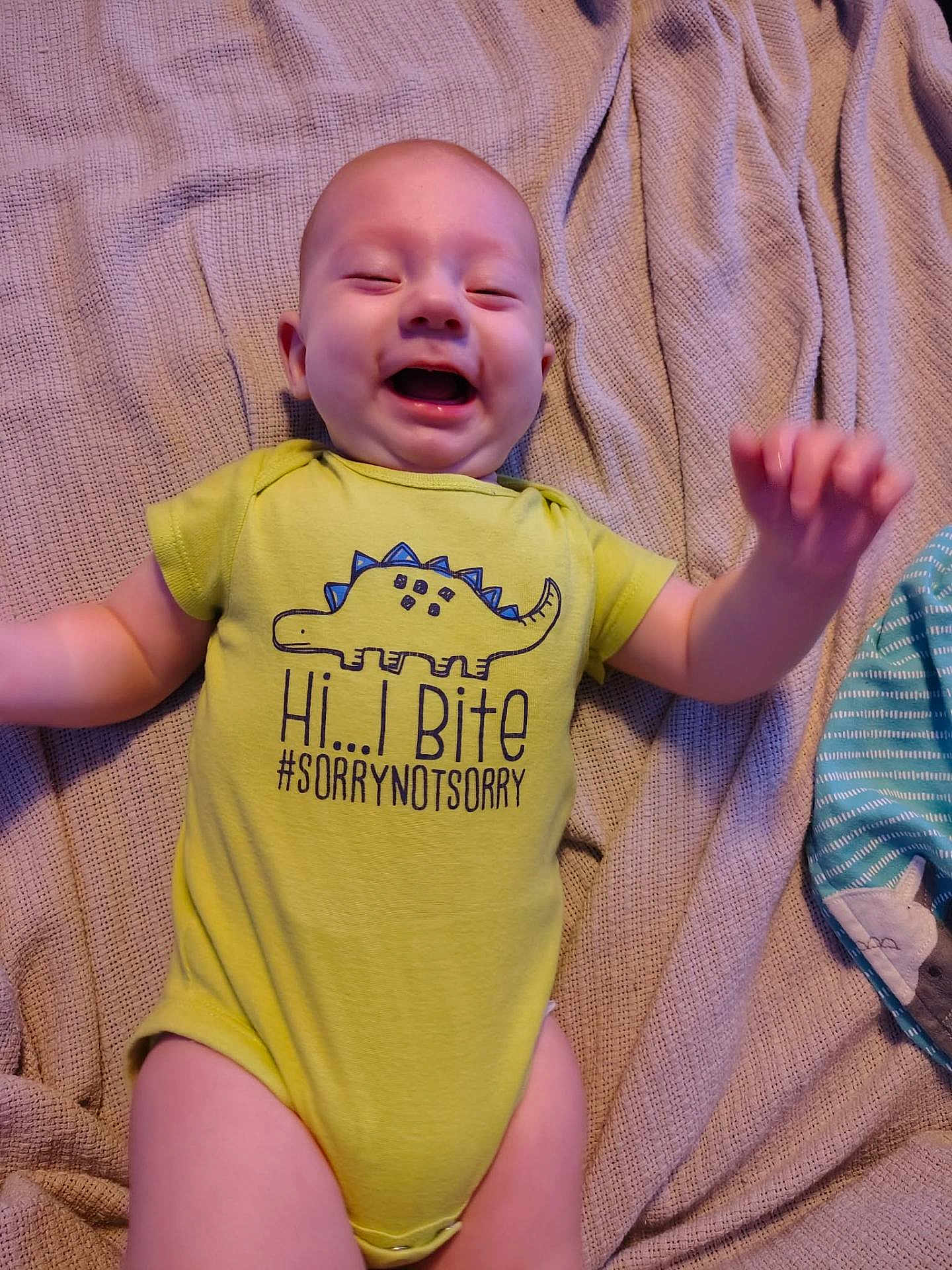 Wyatt is registered to the contest to win money with this photo: baby, infant, smiling, laughing, yellow_onesie, dinosaur_print, text_on_clothing, blanket, lying_down, happy, cute, child, toddler, indoors, fabric, clothing, person, face, head, joyful