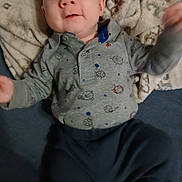 Wyatt is registered to the contest to win money with this photo: baby, infant, child, blanket, clothing, grey_shirt, dark_pants, pattern, lying_down, hand, face, person, cute, indoors, fabric, soft_texture, happy, smile, young, portrait