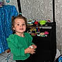 Ana a rejoint le concours — aidez-le/la à gagner de superbes lots ! toddler, child, green_clothing, barefoot, indoor, toy, play_stand, striped_canopy, tile_floor, wallpaper, black_furniture, colorful_toys, person, young_child, smiling, curly_hair, holding_object, home, play_area, standing
