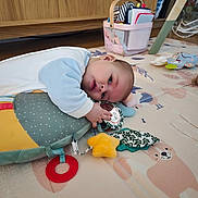 Soalina a rejoint le concours — aidez-le/la à gagner de superbes lots ! baby, infant, playmat, pillow, toy, plush_toy, teether_ring, yellow_star, hand, face, eyes, sleep_suit, wooden_cabinet, storage_basket, books, floor, soft_toys, childroom, laying_down, closeup