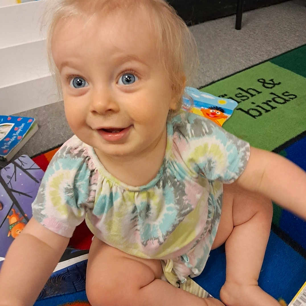 Avree is registered to the contest to win money with this photo: baby, blue_eyes, books, carpet, child, colorful, curious, cute, early_learning, floor, happy, indoor, infant, person, play_mat, sitting, smiling, tie_dye_shirt, toys, young