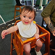 Isla joined the competition — help win amazing prizes! baby, high_chair, bib, smiling, child, wooden_furniture, tattoo, outdoor, casual, happy, person, footwear, white_shoes, face, arm, chair, floor, green_wall, daylight, reaching_hand