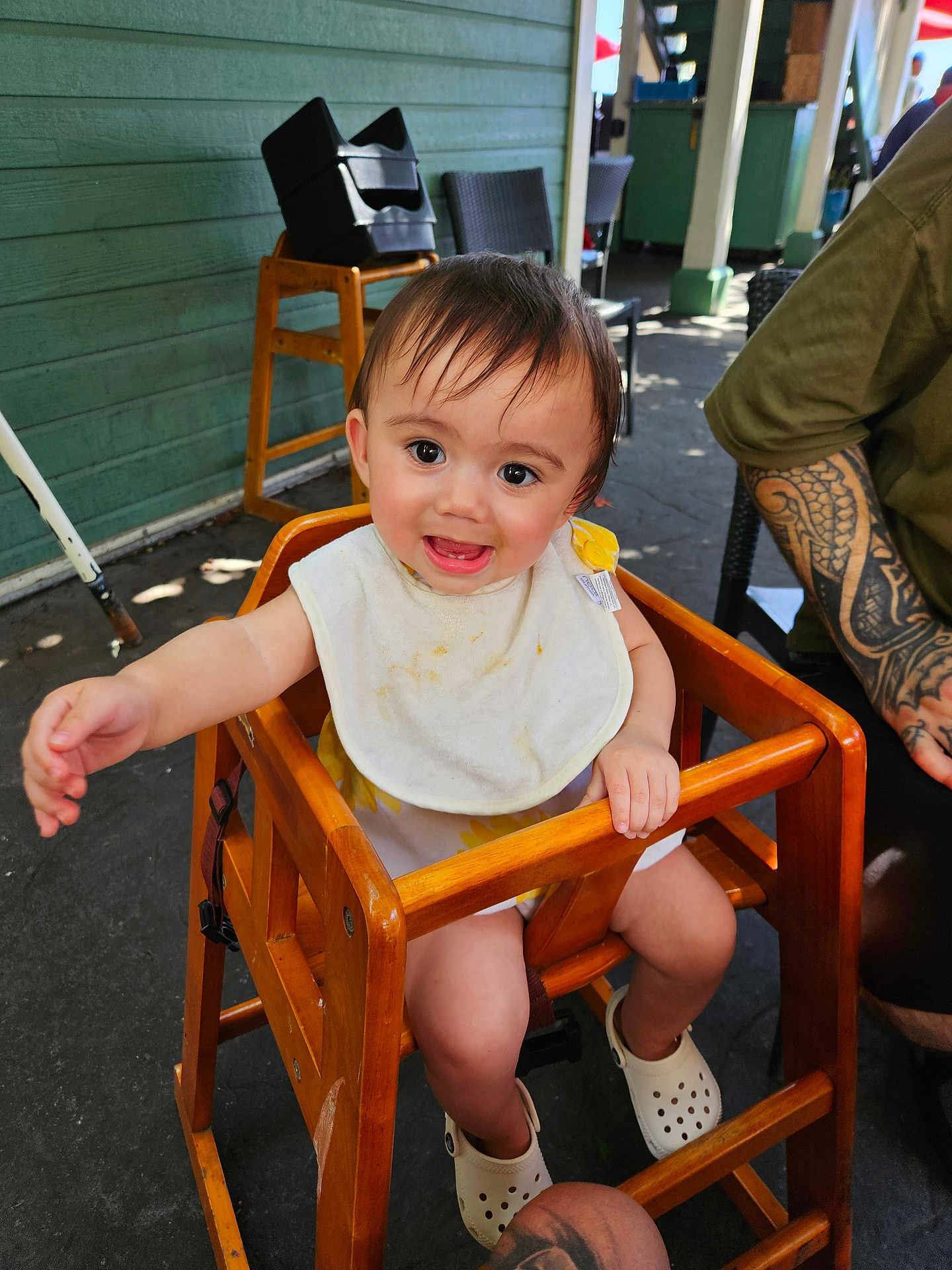 Isla joined the competition — help win amazing prizes! baby, high_chair, bib, smiling, child, wooden_furniture, tattoo, outdoor, casual, happy, person, footwear, white_shoes, face, arm, chair, floor, green_wall, daylight, reaching_hand