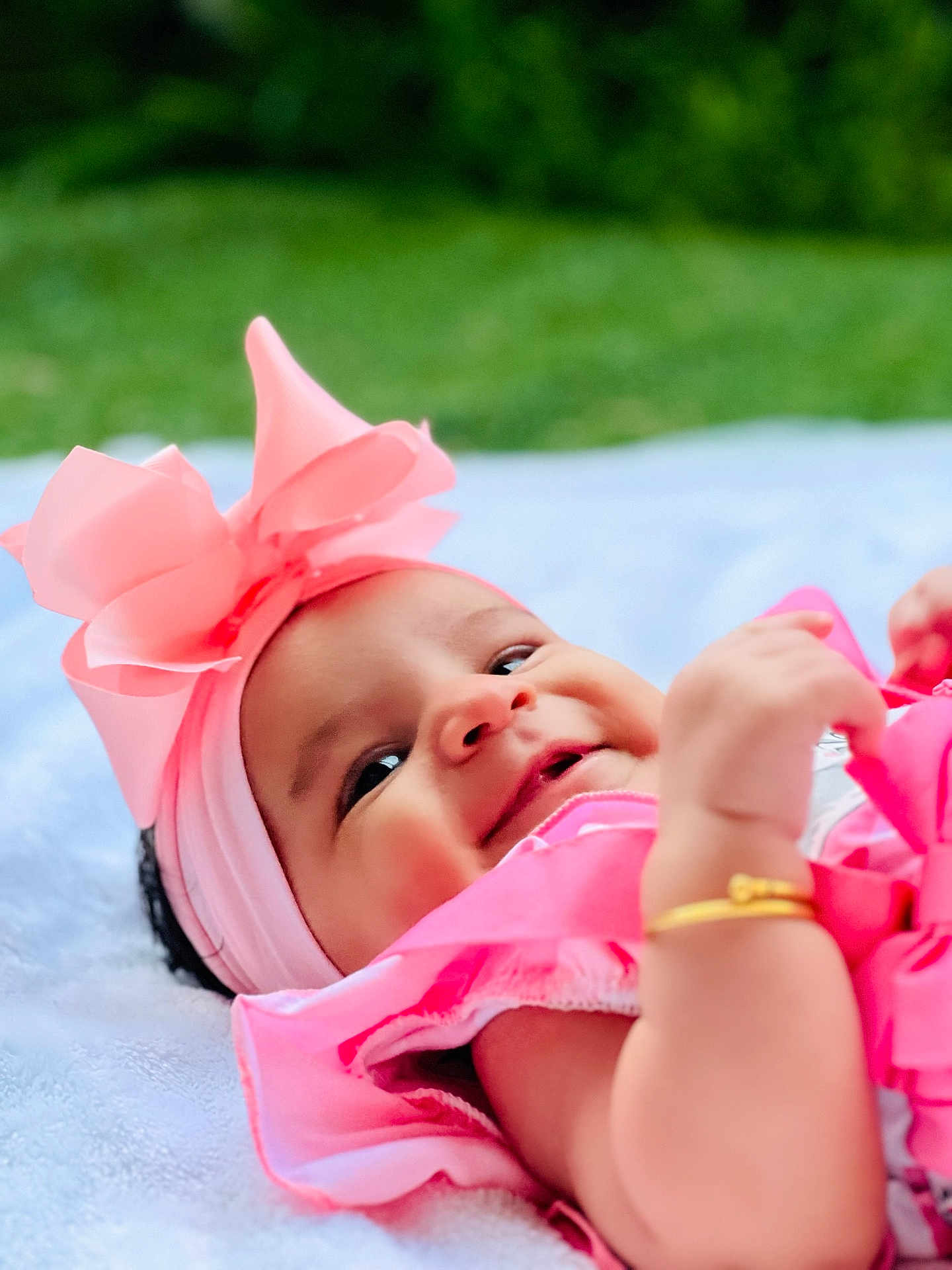 Aaina joined the competition — help win amazing prizes! baby, pink_bow, headband, smiling, outdoor, green_background, blanket, golden_bracelet, infant, cute, happy, portrait, child, soft_texture, closeup, baby_clothing, toddler, playful, young_child, natural_light