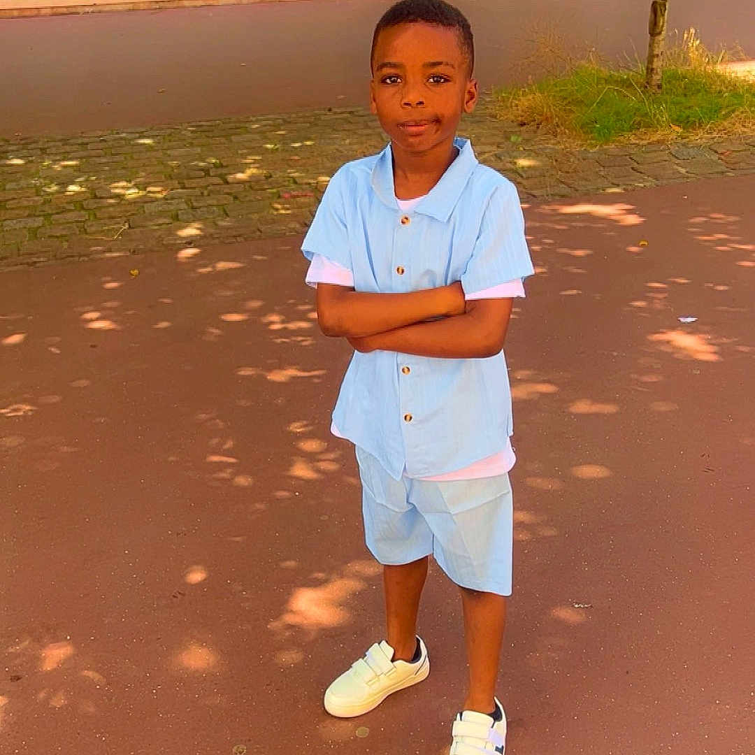 Yohann Nolan participe au concours pour gagner de l'argent avec cette photo : arms_crossed, blue_shirt, boy, building, casual_clothing, child, confident, outdoor, pavement, person, shadow, shorts, standing, street, summer, sunlight, tree, white_sneakers, window, young