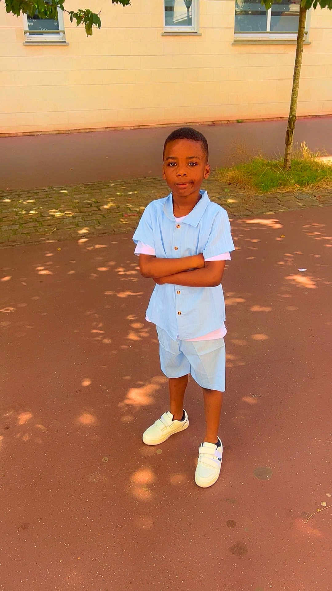 Yohann Nolan participe au concours pour gagner de l'argent avec cette photo : child, boy, standing, outdoor, casual_clothing, blue_shirt, shorts, white_sneakers, tree, pavement, shadow, building, window, sunlight, confident, arms_crossed, young, person, summer, street