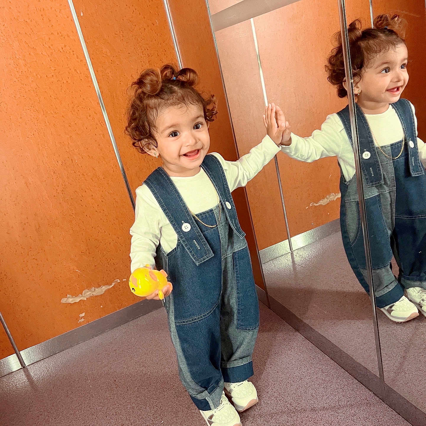 Duaa is registered to the contest to win money with this photo: casual_clothing, child, curly_hair, cute, denim_overalls, floor, hand_touching, happy, indoor, mirror, orange_wall, person, playful, reflection, shoes, smiling, standing, toddler, toy, young_child