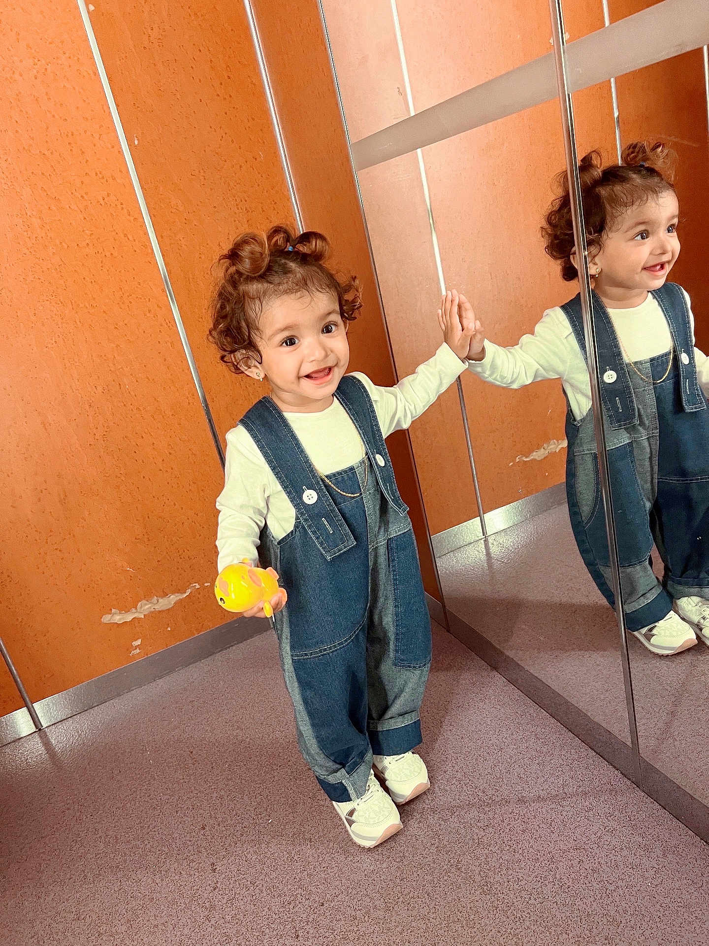 Duaa is registered to the contest to win money with this photo: child, toddler, smiling, curly_hair, denim_overalls, toy, mirror, reflection, orange_wall, floor, shoes, happy, indoor, person, playful, hand_touching, casual_clothing, cute, young_child, standing