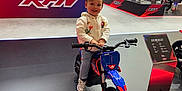 Sebastian a rejoint le concours — aidez-le/la à gagner de superbes lots ! child, toddler, smiling_child, mini_motorbike, blue_motorbike, trade_show, exhibition, display_booth, rfn_logo, platform, handlebars, wheel, hoodie, jeans, sneakers, indoor, people, motorcycles, crowd, floor