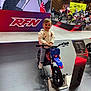 child, toddler, smiling_child, mini_motorbike, blue_motorbike, trade_show, exhibition, display_booth, rfn_logo, platform, handlebars, wheel, hoodie, jeans, sneakers, indoor, people, motorcycles, crowd, floor