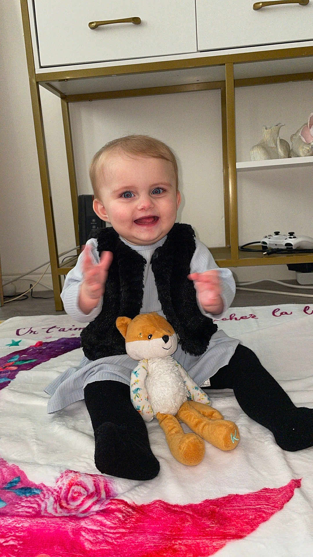 Isalya participe au concours pour gagner de l'argent avec cette photo : baby, child, smiling, clapping, plush_toy, fox_toy, blanket, black_socks, gray_dress, black_vest, indoor, floor, furniture, shelf, game_controller, happy, cute, portrait, person, toddler