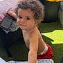baby, bare_chest, blanket, candid, child, curly_hair, diaper, expression, face, floral_pattern, furniture, green_carpet, hand, outdoor, playful, red_shorts, smartphone, sunlight, toddler, wicker_chair