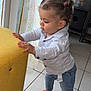 toddler, child, indoor, curly_hair, pigtails, barefoot, yellow_ottoman, white_shirt, blue_jeans, tile_floor, curtains, natural_light, standing, leaning, cute, young_child, home, casual_clothing, side_view, small_hands