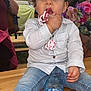 toddler, child, pacifier, curly_hair, jeans, shirt, water_bottle, table, flower_arrangement, indoor, person, face, hand, foot, sitting, casual_clothing, curious, brown_eyes, skin, background