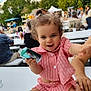 casual, child, crowd, curious, daylight, event, hair_buns, happy, holding_object, nature, outdoor, people, person, pink_outfit, playful, shorts, smiling, table, toddler, trees