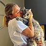 woman, cat, hugging, indoor, blonde_hair, white_shirt, striped_pants, tattoo, bracelets, green_eyes, fluffy_fur, affection, pet, love, portrait, casual_clothing, home, furniture, natural_light, cozy