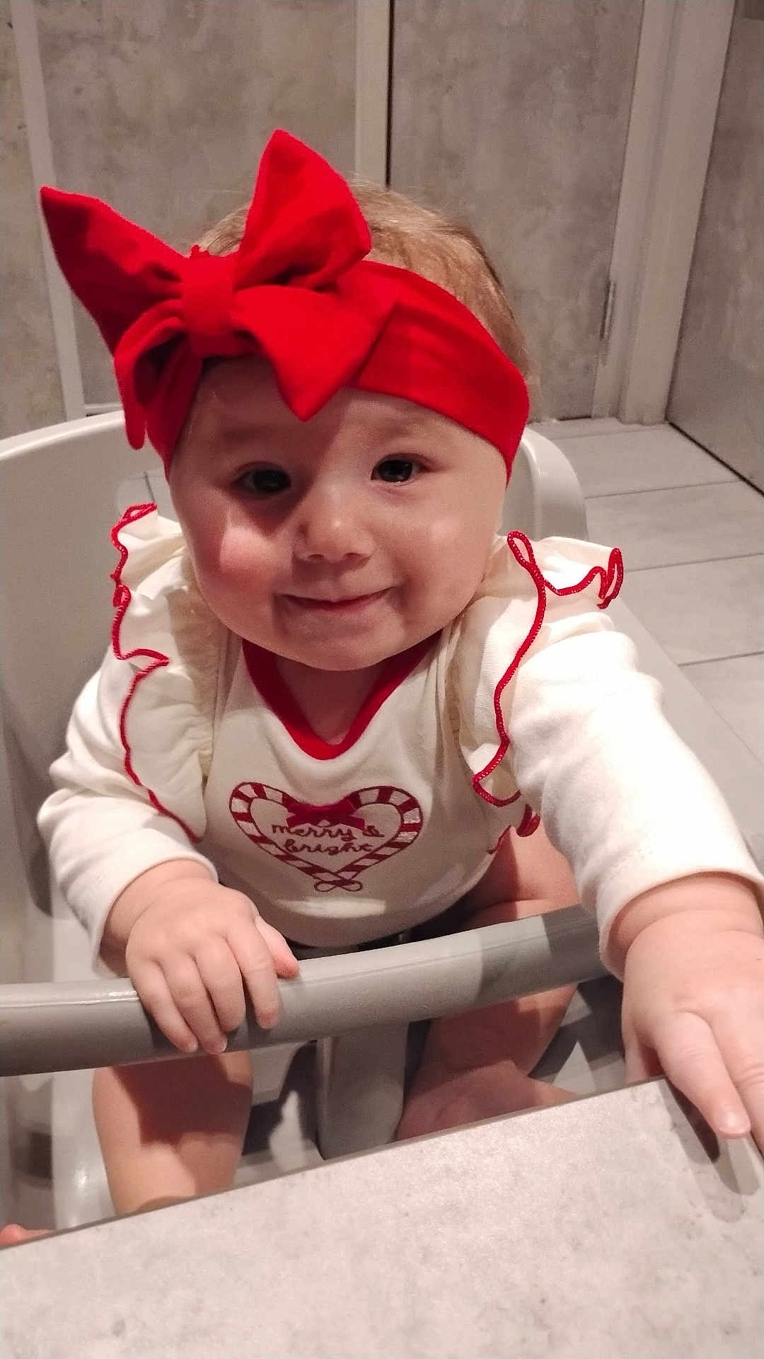 Violet is registered to the contest to win money with this photo: child, baby, highchair, red_headband, bow, smile, face, cheeks, hands, onesie, ruffles, heart_graphic, indoor, tiled_wall, floor, tray, seat, portrait, cute, holiday_outfit