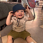 Andréas a rejoint le concours — aidez-le/la à gagner de superbes lots ! toddler, child, smiling, hat, bucket_hat, indoor, floor, tiled_floor, sneakers, shorts, shirt, baby_bottle, sofa, high_chair, living_room, happy, person, footwear, casual_clothing, baby