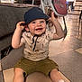 toddler, child, smiling, hat, bucket_hat, indoor, floor, tiled_floor, sneakers, shorts, shirt, baby_bottle, sofa, high_chair, living_room, happy, person, footwear, casual_clothing, baby